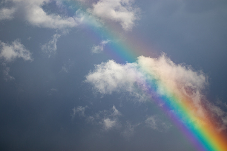 Image of a rainbow showing in a blue sky between white whisps of clouds.